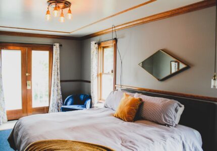 A bedroom with a large bed featuring white and gray bedding, a mustard-yellow throw pillow, and a blanket. There is a window with floral curtains, a blue armchair in the corner, a pendant light, and a diamond-shaped mirror on the wall.