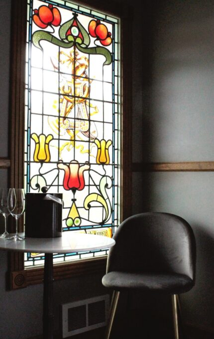 A room with a stained glass window featuring a colorful pattern. Next to the window is a small round table with two wine glasses and a dark container. Two gray chairs are positioned on either side of the table. Floral curtains cover part of another window.