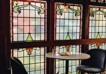 The image shows a small round table with two dark-colored chairs in front of three large stained glass windows with intricate designs. There is a single hanging light bulb above the table, which has a few items placed on it. The setting appears cozy and intimate.