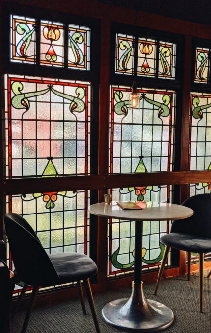 The image shows a small round table with two dark-colored chairs in front of three large stained glass windows with intricate designs. There is a single hanging light bulb above the table, which has a few items placed on it. The setting appears cozy and intimate.