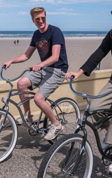 A young man and woman are riding bicycles along a path by the beach. The man is wearing a black t-shirt and shorts, while the woman is wearing a black sweater and pants. They are smiling and seem to be enjoying the sunny day. The beach and ocean are in the background.