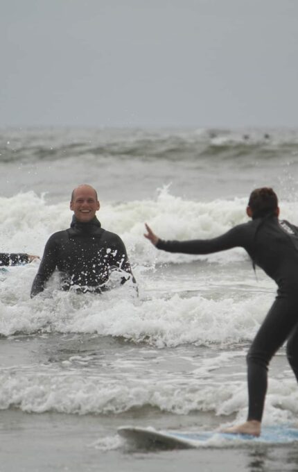 Three people are surfing near the shore with waves breaking around them. One person is standing on a surfboard, another is sitting on a surfboard, and the third is in the water smiling. All are wearing wetsuits. The sky is overcast.