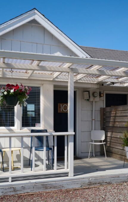 A small white house with a wooden pergola covering the porch. The porch has a few chairs, and a hanging basket with flowers. The house has several windows with blinds, two doors, and plants in pots. The sky is clear and blue.