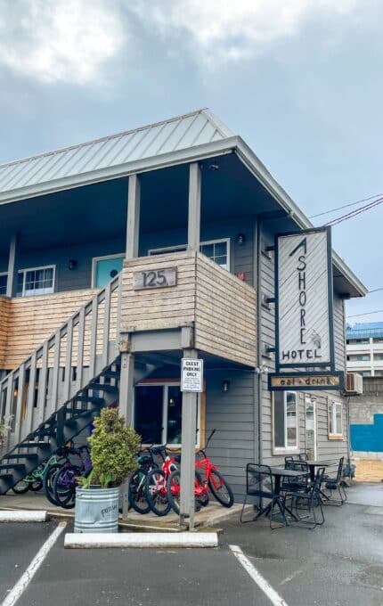 The image shows a two-story building labeled "Shore Hotel" with the building number 125 displayed on a second-floor railing. Bicycles are parked under an outdoor staircase, and a few tables and chairs are set outside. A parking area and cloudy sky are visible.