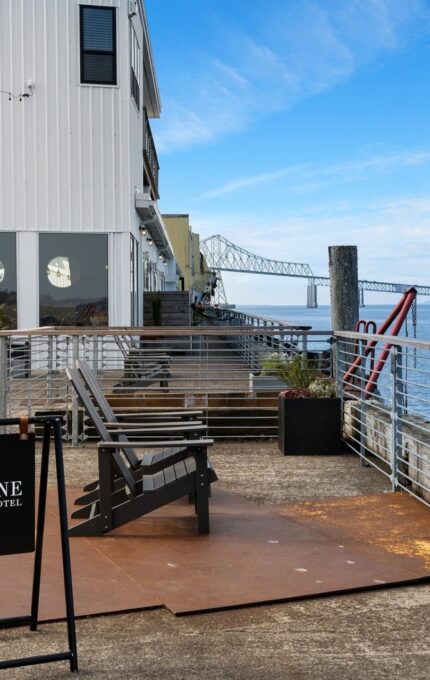 Outdoor patio of Bowline Hotel with seating areas, plants, and a view of a large bridge extending over the water. The patio is fenced with metal railings, and the weather is clear with a blue sky.