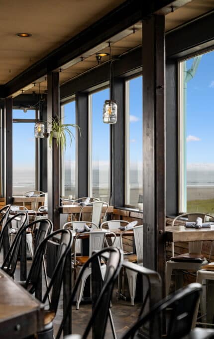 A beachfront restaurant interior with large windows offering an ocean view. Modern, industrial-style seating includes black and white metal chairs. Rolled napkins and glass light fixtures are visible. A plant is hanging near a window. Daylight illuminates the scene.