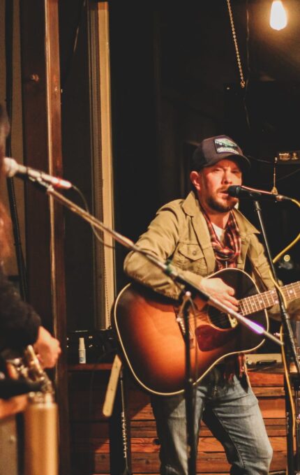 Two musicians perform on stage; one, in the foreground, stands singing and playing an acoustic guitar. The other, partially visible, is also playing an instrument. The setting appears to be an intimate venue, with warm lighting and visible microphones.