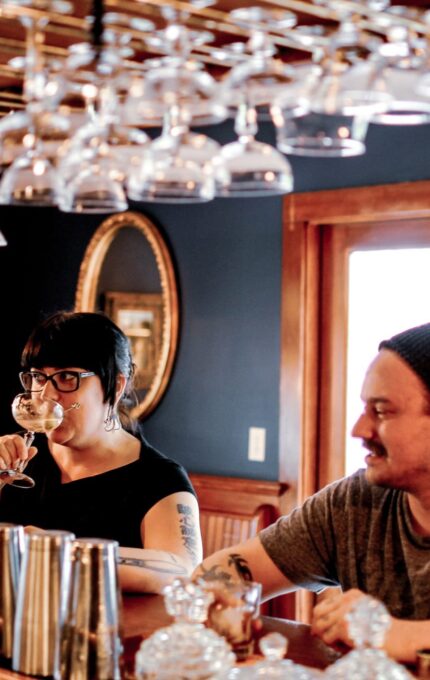 Two people are seated at a wooden bar. The person on the left is drinking from a wine glass, and the person on the right, wearing a beanie, looks ahead. There is a row of upside-down hanging glasses and a stained glass window in the background.