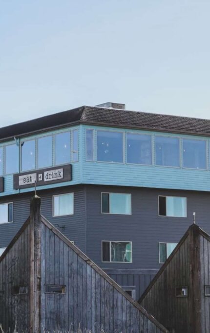 A multi-story building with a blue and gray exterior. The upper level features large windows and signage that reads "eat + drink" and "lodging & spa." In the foreground, there are two smaller wooden structures with triangular roofs. There are trees to the right.