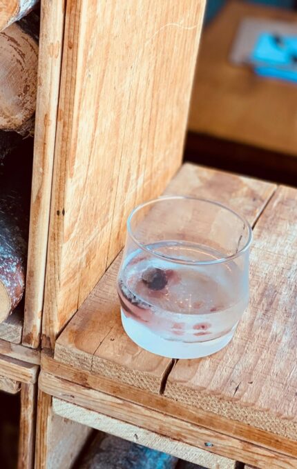 A neatly stacked pile of cut wood logs inside a wooden storage unit, with a clear glass containing a beverage and ice cubes placed on top of the unit. A partially visible blue notebook is in the background.