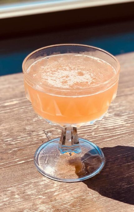 A filled glass with a light orange beverage sits on a wooden table. The cocktail glass has a short stem and a wide bowl, casting a shadow to the right due to the sunlight.