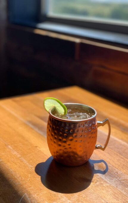 A copper mug filled with a beverage, garnished with a lime slice, is placed on a wooden table by a window with natural light illuminating the scene. The background shows part of the window frame and an out-of-focus outdoor view.