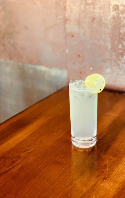 A tall, clear glass filled with a cold, light-colored beverage, garnished with a lemon slice, sits on a polished wooden table. The background features a textured, pinkish wall.