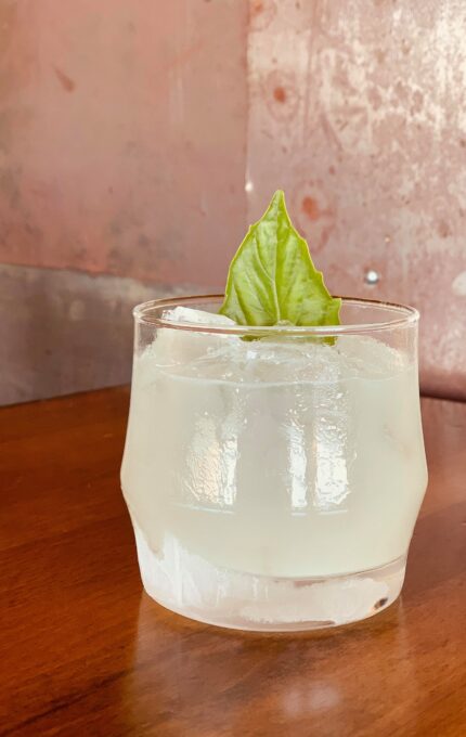 A clear cocktail in a rounded glass with ice and a green leaf garnish sits on a wooden table against a textured, pale pink background.