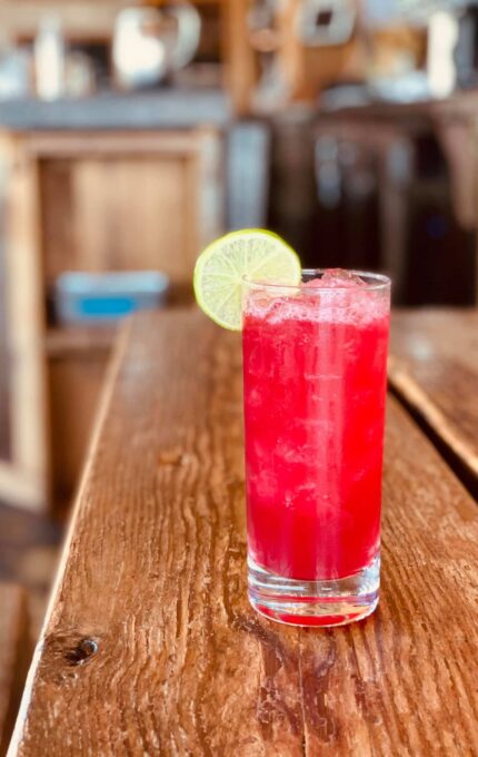 A tall glass filled with a red-colored beverage, topped with ice and garnished with a lime wheel on the rim, placed on a rustic wooden table. The background is slightly blurred, featuring a wooden interior with indistinct objects.