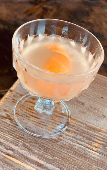 A clear glass goblet filled with a light-colored liquid sits on a wooden surface. An orange peel is floating inside the drink. The background appears to be a dark, rustic wooden table.