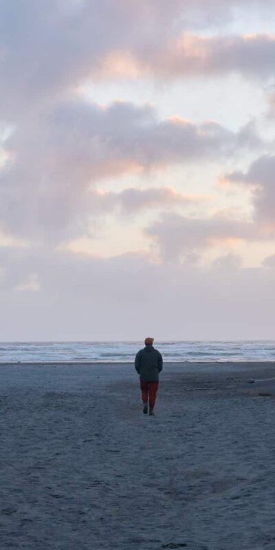 A person walks away from the camera towards the ocean under a cloudy sky during sunset on a sandy beach. Another person is in the distance on the right, also heading towards the water. Grassy dunes border the left side of the image.