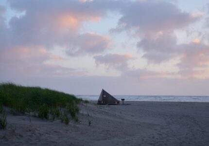 A small wooden pyramid-shaped structure sits alone on a sandy beach with an overcast sky and ocean in the background. Grassy dunes are visible to the left, and the beach is mostly empty. The light from the setting sun casts a soft, pinkish hue on the clouds.