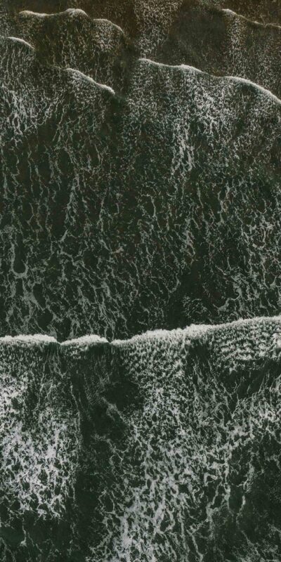 A high-angle view of ocean waves crashing onto the shore. The waves create a pattern of white foam against the dark green water, revealing the texture of the sea's surface.