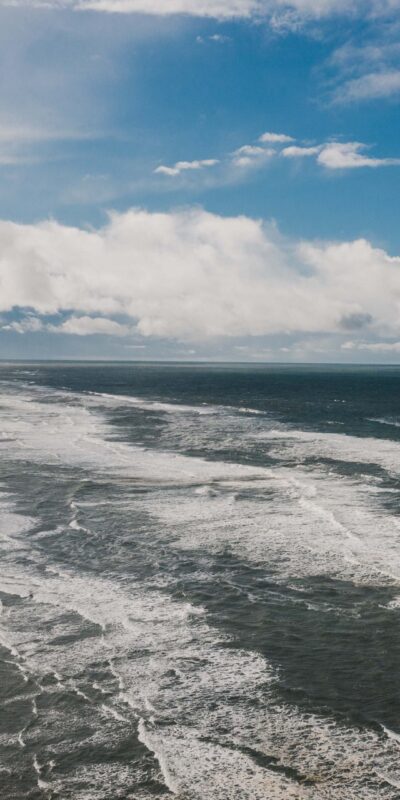 Aerial view of a wide beach with waves crashing onto the shore. The sky is partly cloudy, and the horizon is visible in the distance. Sparse vegetation is seen along the beach edge, and a few people are walking along the shoreline.