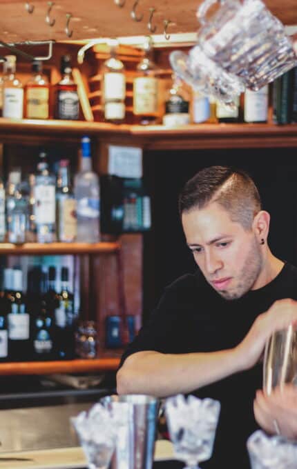 A bartender with short hair is focused on mixing a drink behind a bar. The bar shelves are stocked with various bottles of liquor and wine. There are ice-filled glasses in the foreground, and some glasses are in mid-air, being tossed.