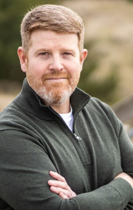 A man with short, reddish-brown hair and a beard, dressed in a dark green zip-up sweater, stands outdoors with his arms crossed. The background is blurred, showing a wooden pathway and natural landscape with greenery and dry grass.