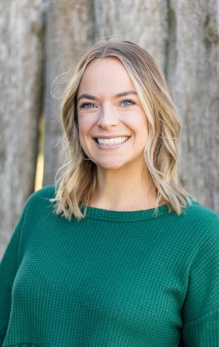 A person with blonde hair and blue eyes is smiling at the camera. They are wearing a green sweater. The background is blurred, featuring a wooden texture.