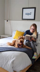 A woman sits on a bed reading a book, with a brown dog lying next to her. The bed has gray and white bedding with a yellow pillow. There is a framed picture of a whale on the wall above the bed. A small bedside table with a hanging light is visible.