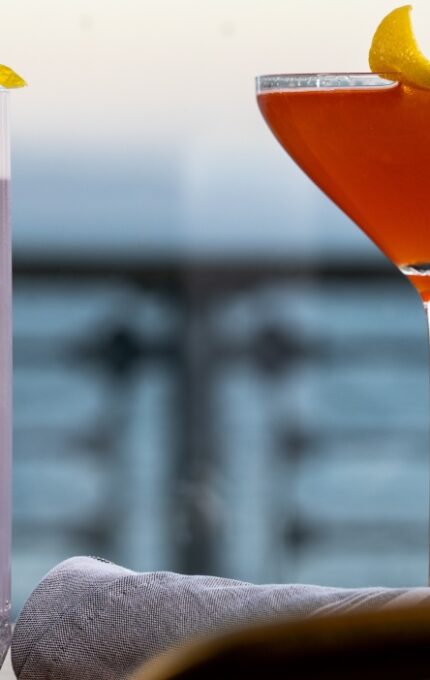 A tall glass of purple cocktail with a foamy top and lemon twist sits next to a martini glass filled with an orange drink, garnished with a lemon twist, on a table by a window overlooking water.