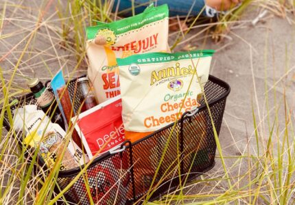 A person sits on the sand surrounded by tall grass, next to a black mesh basket containing various snacks. Visible items include bags of organic cheddar cheese squares, sea salt chips, pretzels, and several bottles.