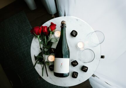 A small round marble table with a bottle of wine, three red roses, four lit candles, two empty wine glasses, and several chocolate pieces arranged neatly. The table is near a white curtain, on a dark floor.