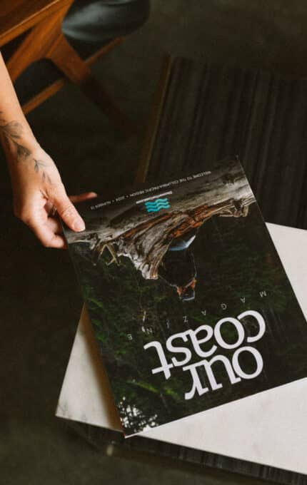 A person with a tattoo on their forearm is holding a magazine titled "Out Coast" over a table. The magazine cover shows an image of a scenic outdoor landscape.