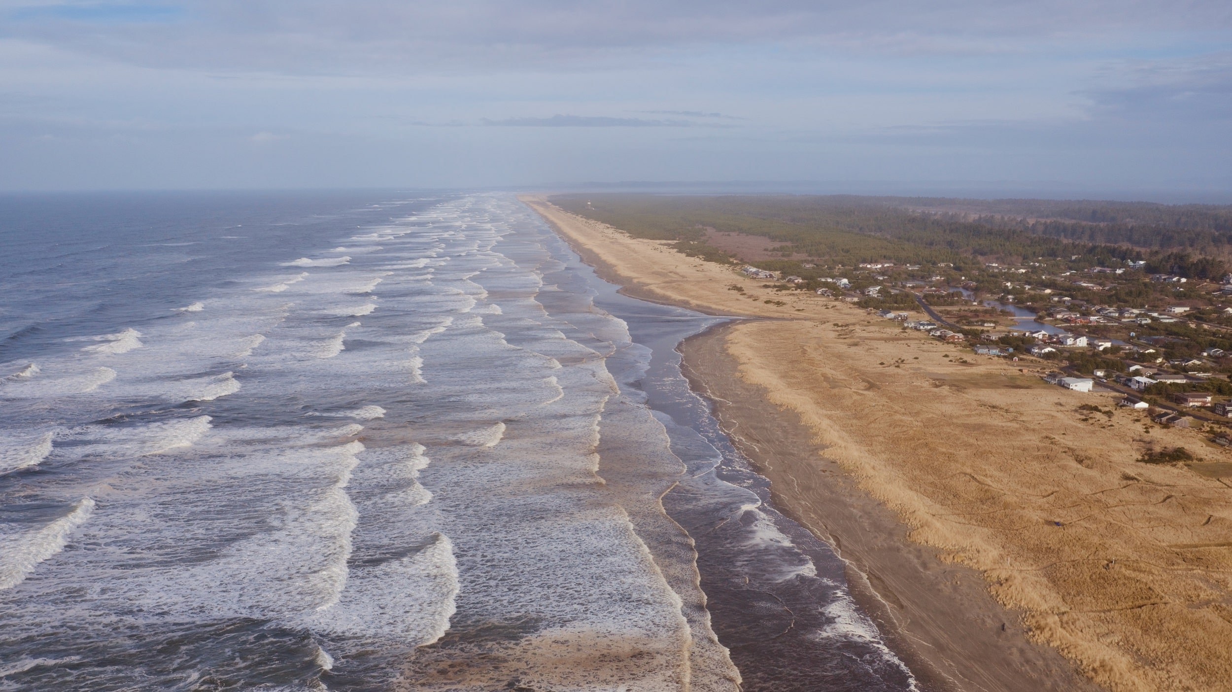 Aerial view of a long sandy beach with gentle waves meeting the shore. On the right, a narrow stretch of dunes separates the beach from a small town. The ocean extends to the horizon, and the sky is partly cloudy.