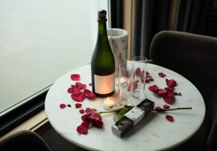 A round marble table with a bottle of sparkling wine, two empty glasses, a candle, rose petals, a single rose, and a box of chocolates next to a window with a view of cloudy or foggy weather.