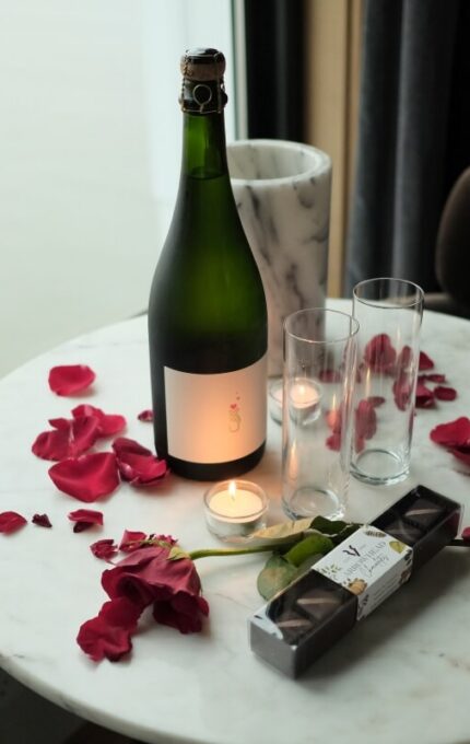 A round marble table with a bottle of sparkling wine, two empty glasses, a candle, rose petals, a single rose, and a box of chocolates next to a window with a view of cloudy or foggy weather.