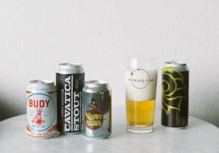 Five cans of craft beer are arranged on a round white table next to a glass of beer with foam, labeled "Pickled Fish." The background is a plain, light-colored wall.
