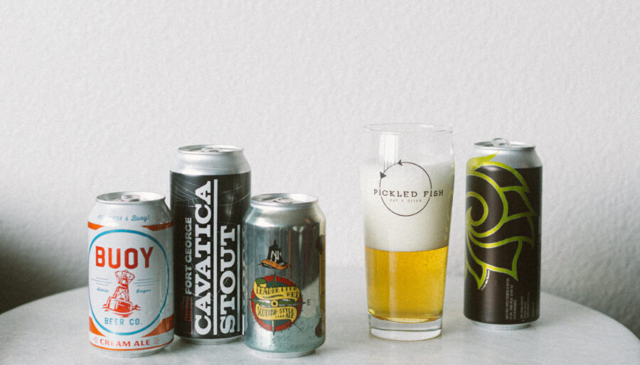 Five cans of craft beer are arranged on a round white table next to a glass of beer with foam, labeled "Pickled Fish." The background is a plain, light-colored wall.