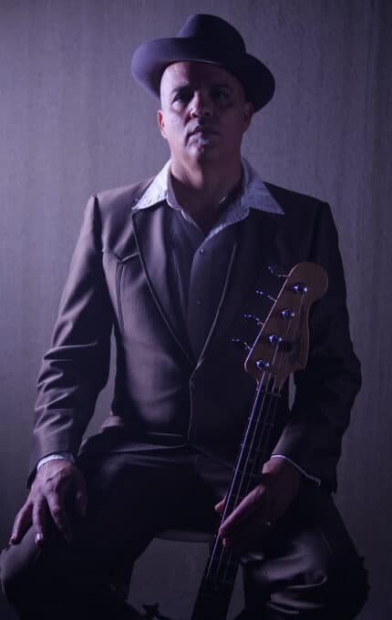 A person wearing a dark suit and hat is seated, holding a bass guitar vertically. The lighting is dim, creating a moody atmosphere. The background is a plain, neutral-colored surface.