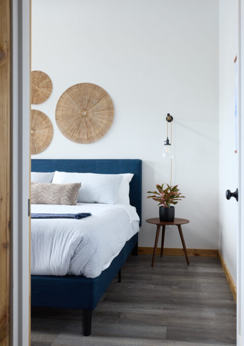 A bedroom with a blue bed, white sheets, and assorted pillows. Three round wicker decorations hang on a white wall. A small wooden side table holds a potted plant under a wall-mounted lamp. The room has wood flooring and a wood door frame.