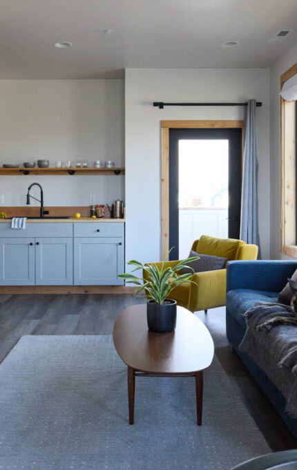 A modern living room with a blue sofa, a black dog lying on it, and a wooden coffee table. A wall-mounted TV and a fireplace are on the left. The kitchenette in the background has a sink, cabinets, and open shelving. Large windows provide natural light.