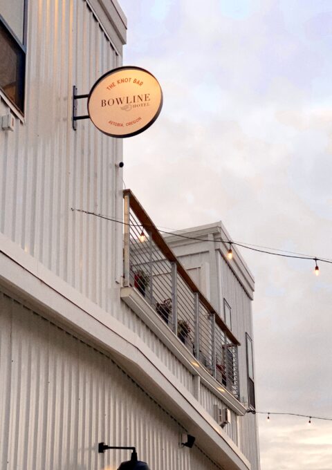 Exterior view of a two-story building with a sign reading "The Knot Bar, Bowline, Astoria, Oregon." A balcony with a railing is visible on the second floor, and string lights hang above. The sky is cloudy.