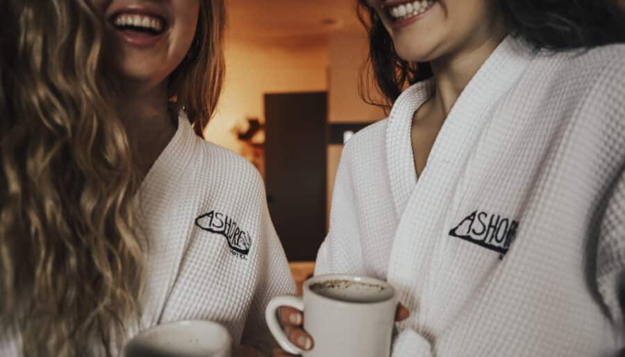 Two people in white robes with embroidered logos hold mugs and smile, standing indoors. The background is softly lit, creating a warm ambiance.