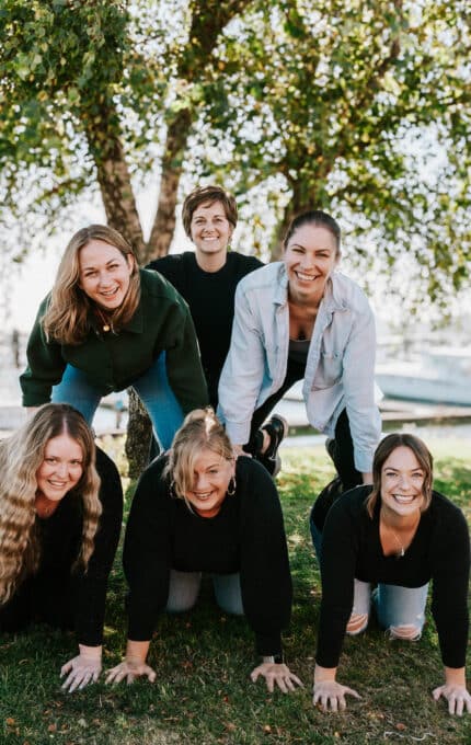 Six people are outdoors, forming a human pyramid on grass with boats and water in the background. Three people are kneeling on the grass, and three others are balanced on their backs, smiling. Trees and sunny weather complete the scene.