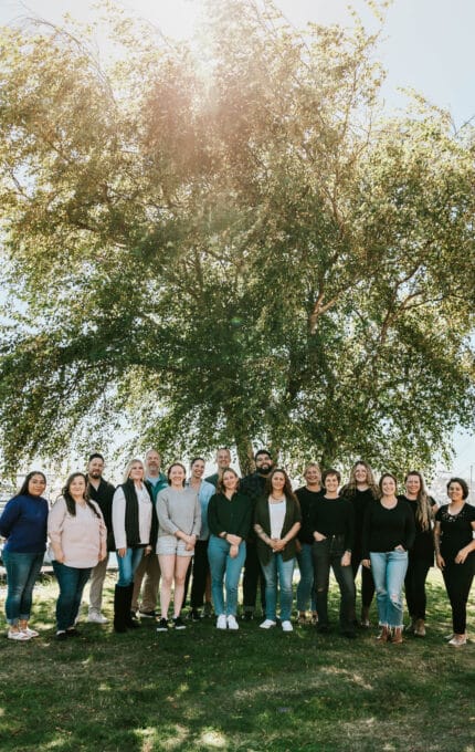 A group of people standing in a semicircle pose for a photo in front of a large tree. It is a sunny day, and the tree provides shade. The group includes both men and women, and they are outdoors on a grassy area.