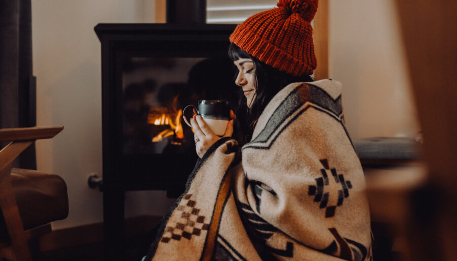 A person sits on the floor wrapped in a patterned blanket, wearing a red knit hat, while holding a mug. They are near a lit wood-burning stove, creating a cozy atmosphere. Wooden furniture and window blinds are visible in the background.