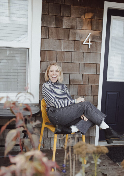 A person sitting on a yellow chair on a porch with crossed legs, smiling. They wear a striped shirt, black jeans, and white shoes. The house has wooden shingles and is numbered 4. There are plants in the foreground and a blue door to the right.
