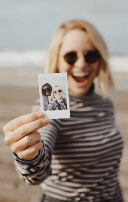 A person wearing sunglasses and a striped shirt holds a Polaroid photo in front of a beach background. The Polaroid shows two people posing together, both wearing sunglasses and smiling.