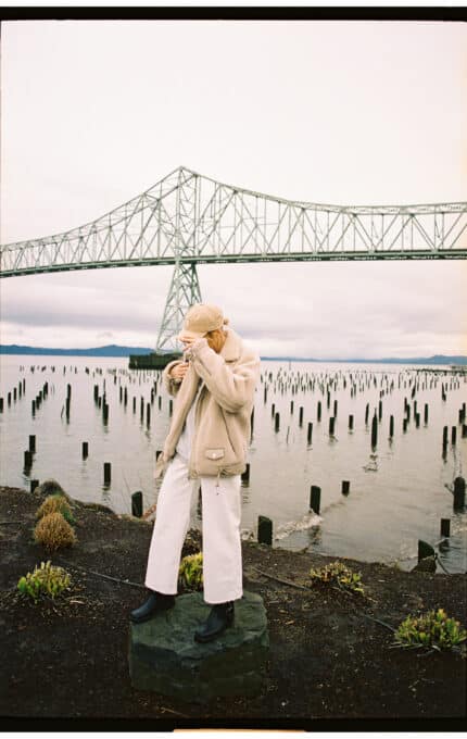 Person wearing a beige jacket and white pants standing on a rock by a body of water. A large bridge spans the background on a cloudy day, with wooden posts rising from the water. The person appears to be adjusting their hat.