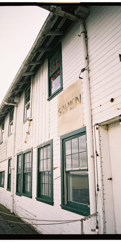 A two-story white wooden building with green-trimmed windows and chipped paint. A sign reads "Salmon For All." String lights hang across the exterior. The sky is overcast.