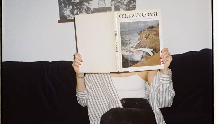 A person sitting on a dark couch holds up a large book titled "Oregon Coast," covering their face. They're wearing a striped shirt, white top, and black pants. A black and white photo of trees and a bridge hangs on the wall behind them.